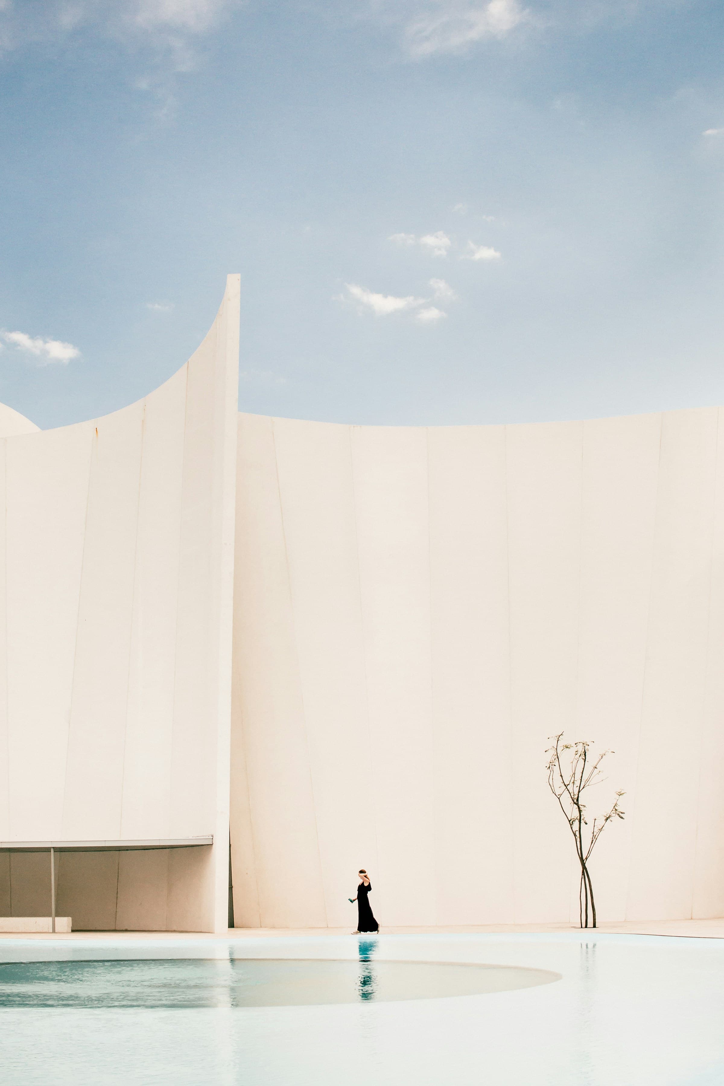 A lone figure walking beside a tall cream curved wall and a still pool